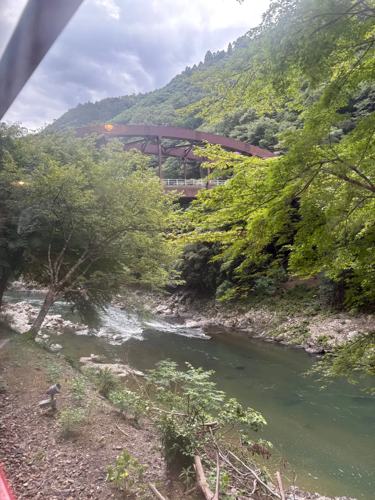 緑豊かな山々と川の風景。中央には赤い鉄製のアーチ橋がかかり、水面は緑色を帯びている。空は曇り、雨が降りそうな様子。