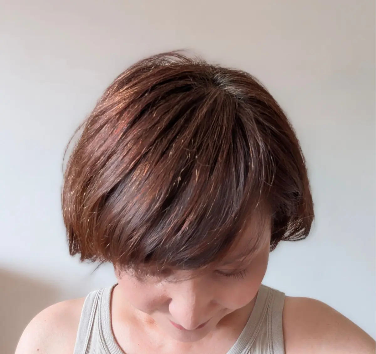 茶色のボブヘアの女性が、前を向いて見下ろしている写真。白い壁の前で、ベージュのタンクトップを着ている。