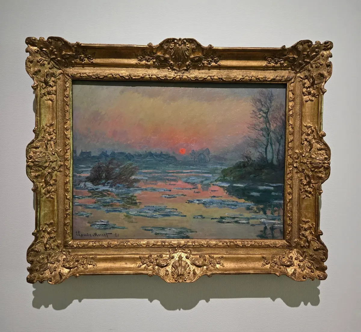 クロード・モネ作の風景画。水面が氷で覆われ、夕焼けのオレンジ色とピンク色が映り込んでいる。右側に木のシルエットが描かれ、金色の装飾的な額縁に入っている。