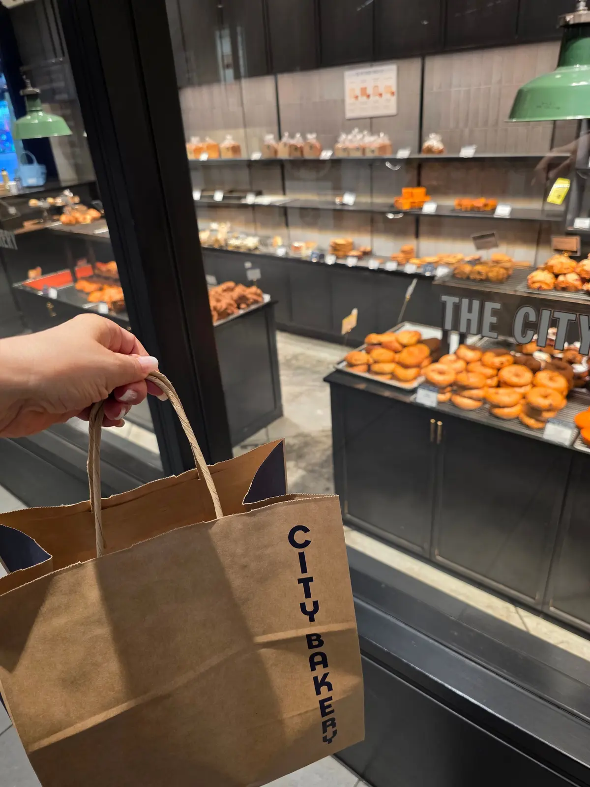 手で提げられた茶色の紙袋に「CITY BAKERY」の文字が印刷されている。背景には、ガラス窓越しにパンが並んだベーカリーの様子が見える。
