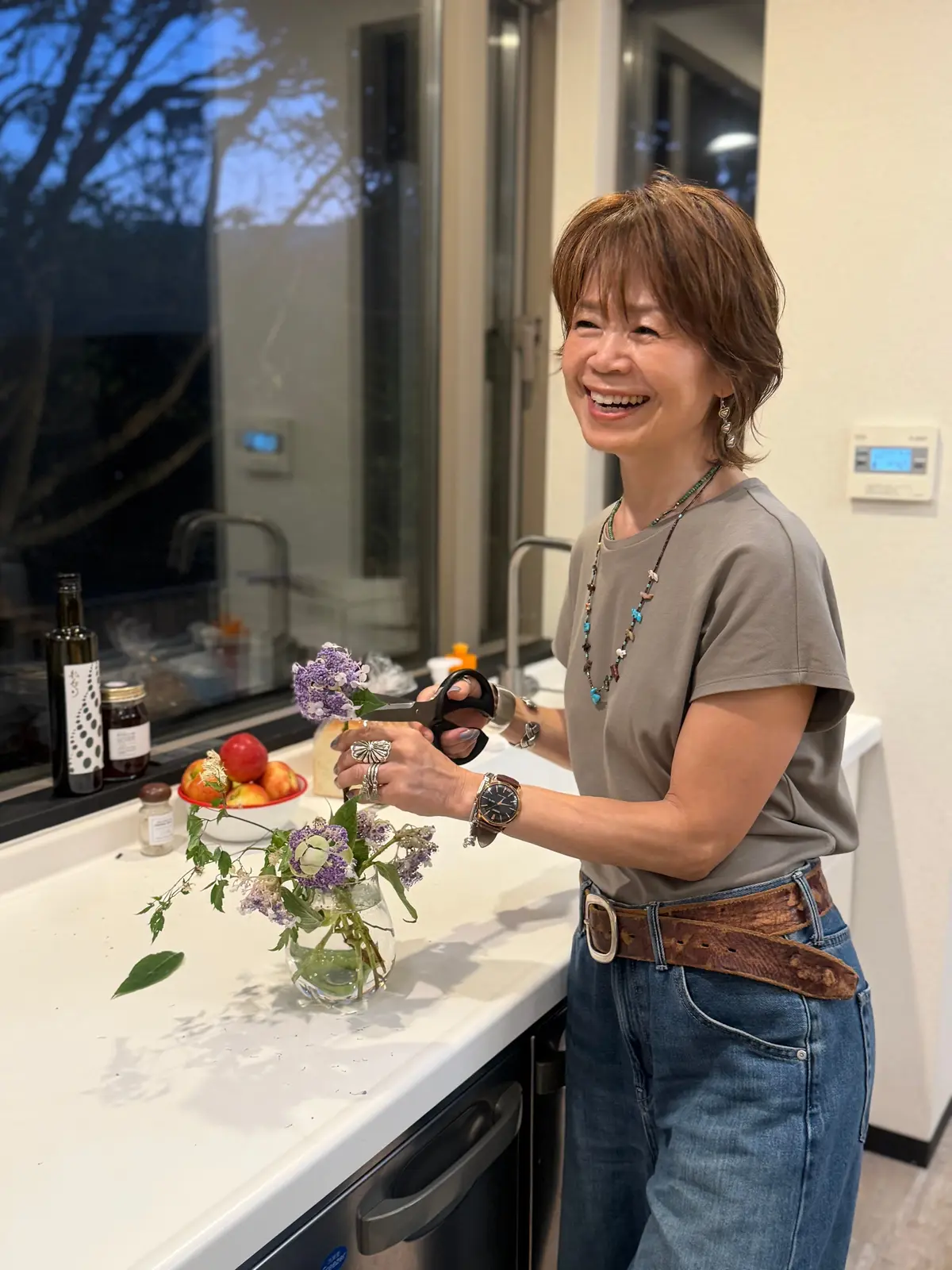 笑顔の女性が、キッチンで小さな花束の手入れをしている。グレーのTシャツにデニム、太めのベルトを着用。花束は透明な瓶に入り、ハサミを持っている。背景には窓とキッチン家電が見える。