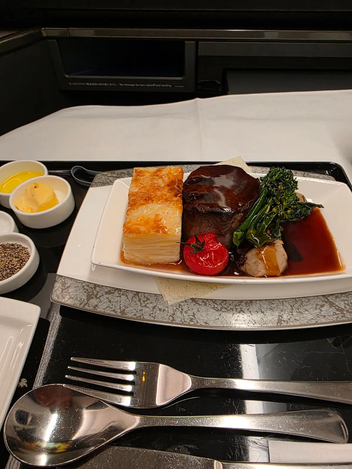 航空機内での食事。白いトレイに、牛肉、マッシュポテト、ブロッコリー、ミニトマトが盛り付けられている。ソースがかかり、調味料の小瓶とカトラリーが添えられている。