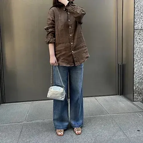茶色の長袖シャツとデニムパンツを着た女性が、銀色のショルダーバッグを持ち、建物のエントランス前で携帯電話に話している。足元にはサンダルが履かれている。