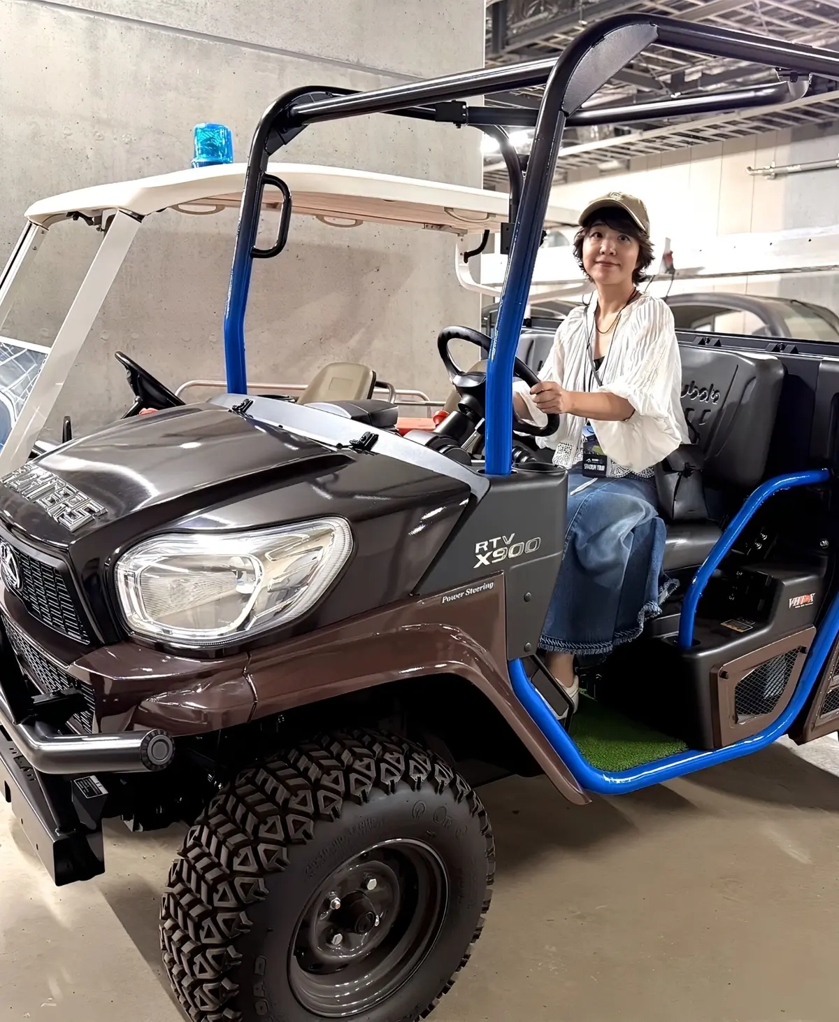 茶色の大型UTVに座る女性。運転席に座り、ハンドルを握っている。白いシャツに青いジーンズ姿。背景はコンクリートの壁。