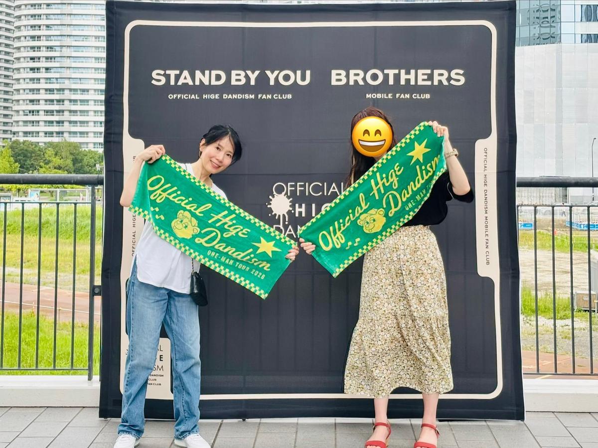 Hige Dandismのファンクラブイベント会場で、若い女性が笑顔でポーズをとっている。緑色の旗を2本持ち、白いトップスとパンツを着用。背景には「STAND BY YOU BROTHERS」のロゴと、テニスコートが見える。