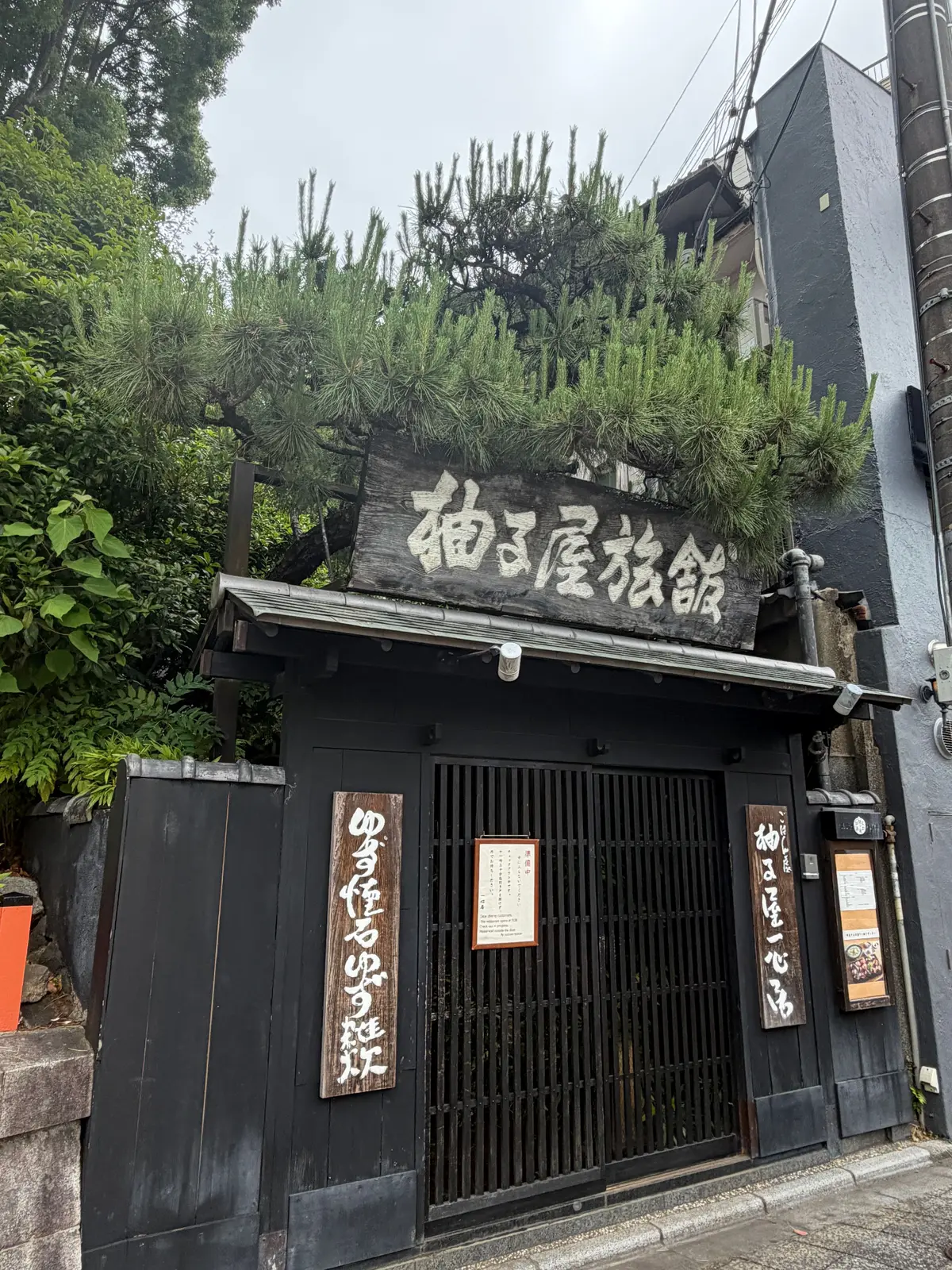 黒い建物の外観で、木製の看板に「和食店」と書かれている。緑の低木が屋根に生い茂り、木製のメニュー表がドアの横に掲げられている。