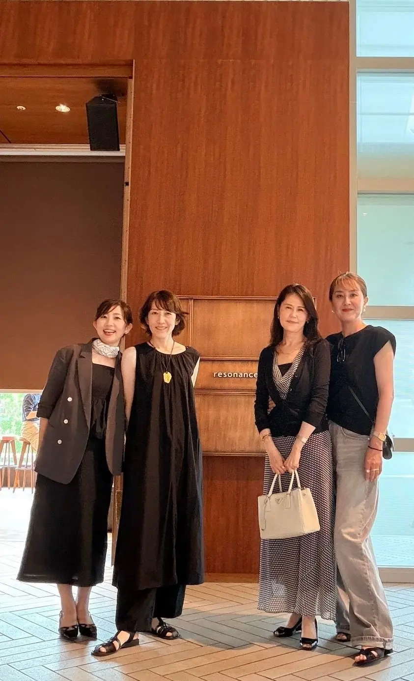 木目調の壁の前に4人の女性が立っている写真。左から黒いジャケットと白い襟付きのトップス、黒いワンピース、水玉模様のトップスとスカート、黒いタンクトップとグレーのパンツを着用。それぞれ異なる表情でカメラを見ている。