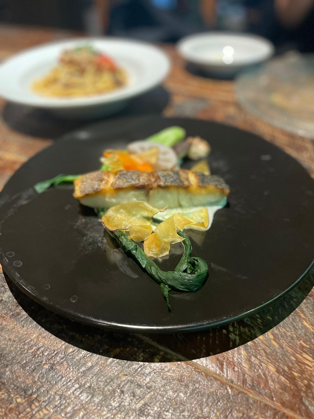 黒い円盤に盛り付けられた魚料理。魚の切り身に、野菜、黄色い葉物、薄い揚げ物が添えられている。背景には、白い皿に入ったパスタ料理が見える。