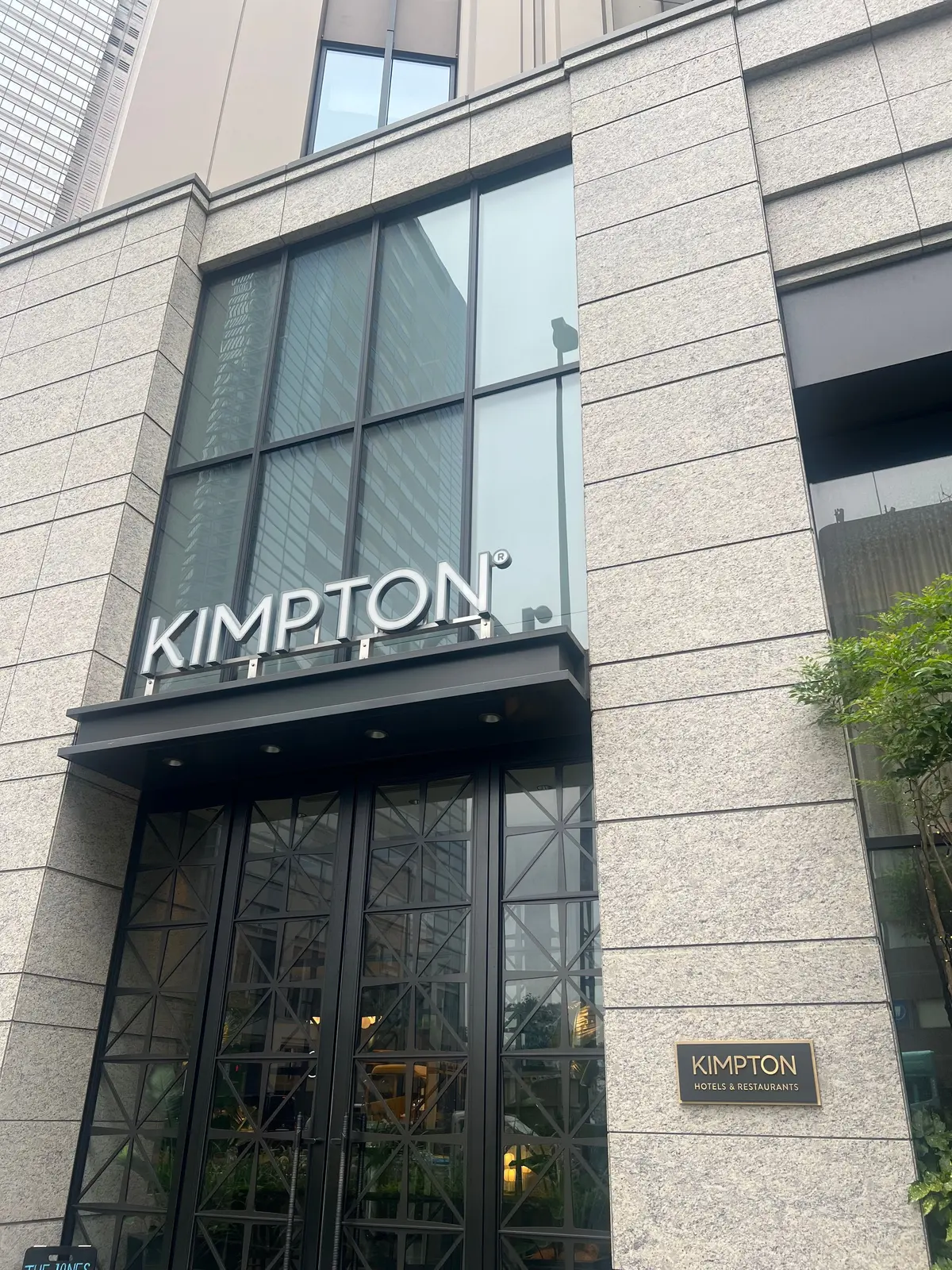 グレーの建物に設置された「KIMPTON」のロゴと入り口。黒いフレームの大きな窓と、右下には「KIMPTON HOTELS & RESTAURANTS」のロゴがある。