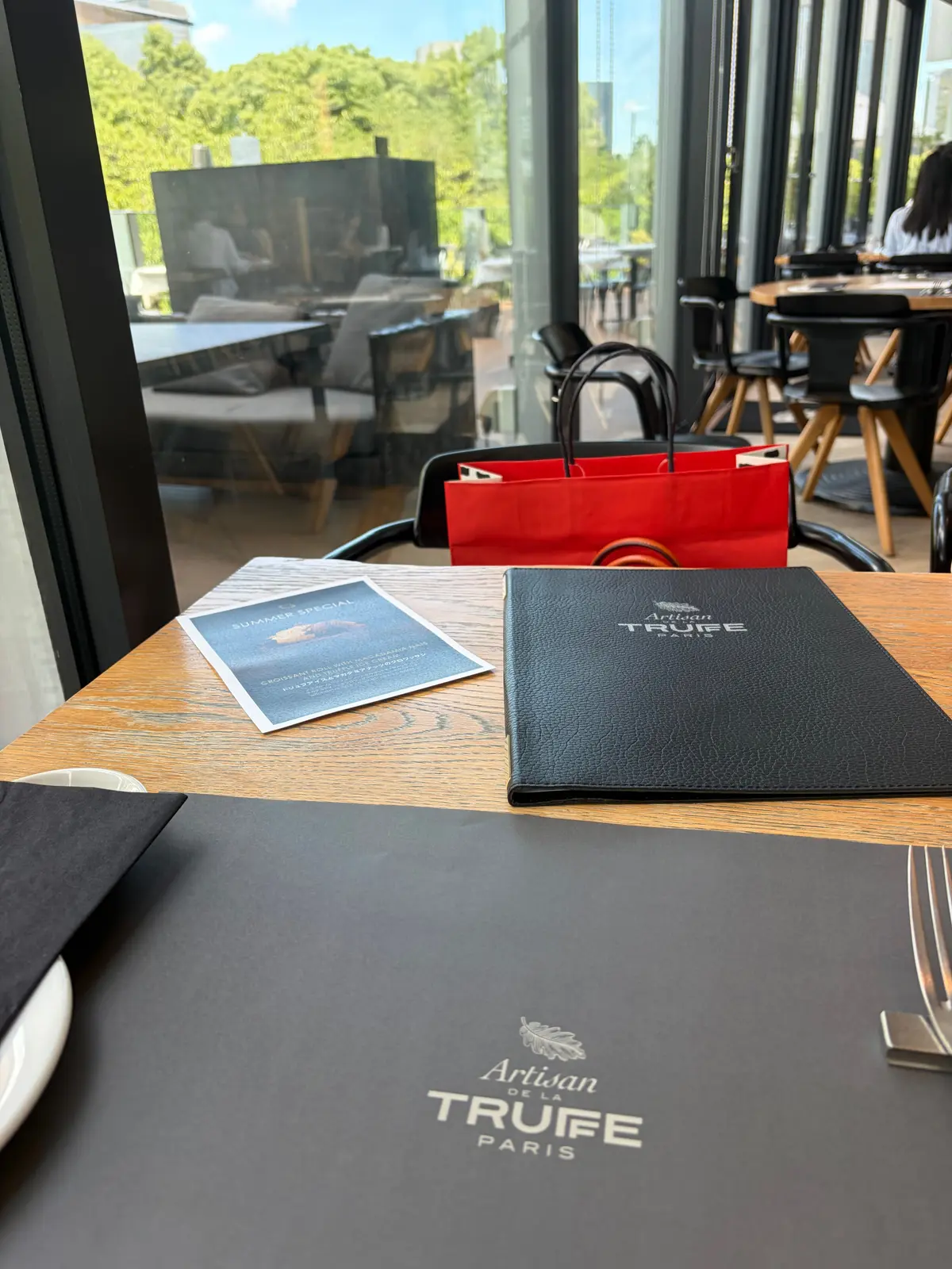 木目調のテーブルに、黒いメニューと「SUMMER SPECIAL」と書かれたチラシが置かれている。テーブルには「Artisan de la Truffe Paris」のロゴが入ったプレースマットがあり、右には赤い紙袋が置かれている。