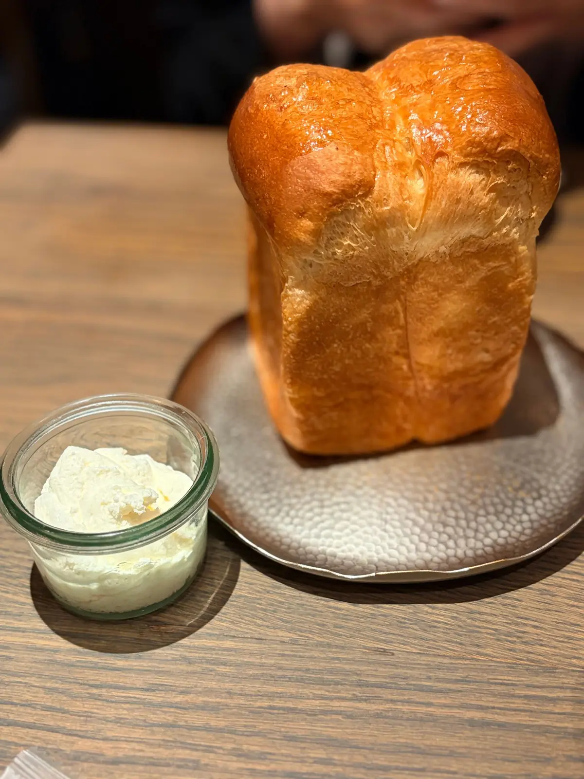 木製のテーブルに置かれた、焼き上げられたパンとバターのセット。パンはふっくらとしたブリオッシュのような形。バターはガラスの器に入っており、パンと一緒に提供されている。