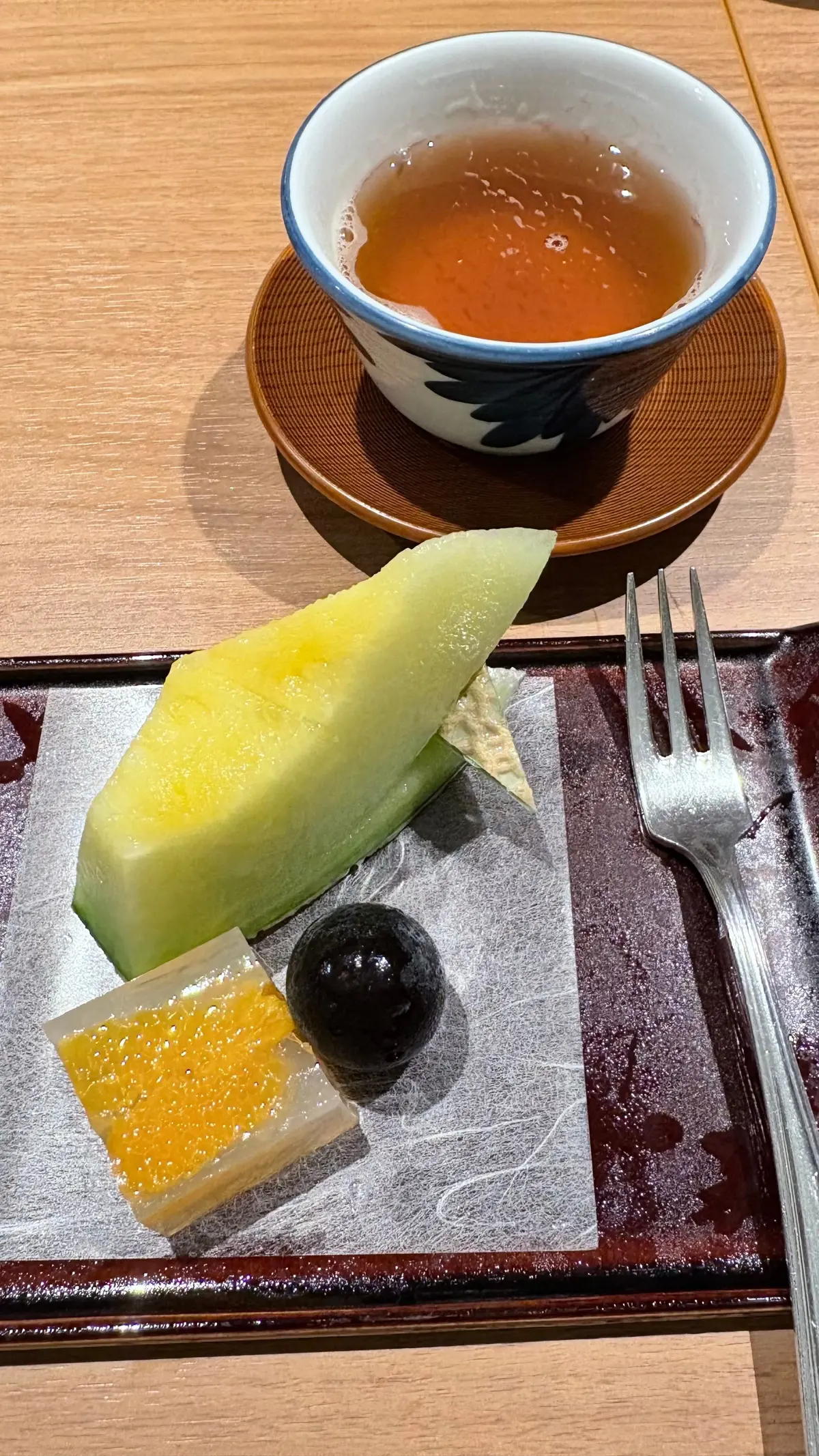 木製のテーブルに置かれた、茶碗に入ったお茶と、和風の盛り合わせ。盛り合わせには、メロン、ゼリー、栗が乗っており、フォークが添えられている。
