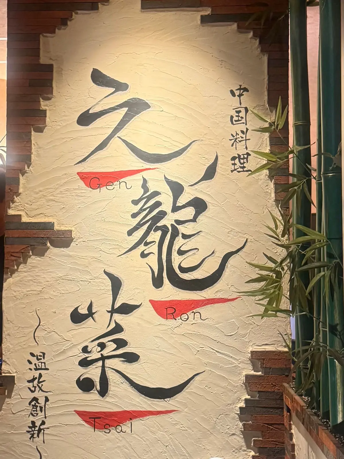 ベージュ色の壁に黒い墨絵風の文字が描かれた装飾。中央に「龍」の文字、左右にそれぞれ中国語の文字が配置。下部には「Gen」「Ron」「Tsai」の文字が赤色の四角形で示されている。壁の左右にはレンガと竹が生えている。