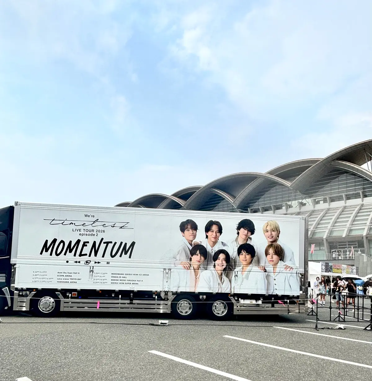 白いトラックの側面には、We're timelesz LIVE TOUR 2026 "MOMENTUM" とツアー情報が大きく表示されている。トラックの前方には、男性と女性のアーティスト写真が配置されている。背景には空港の建物が見える。