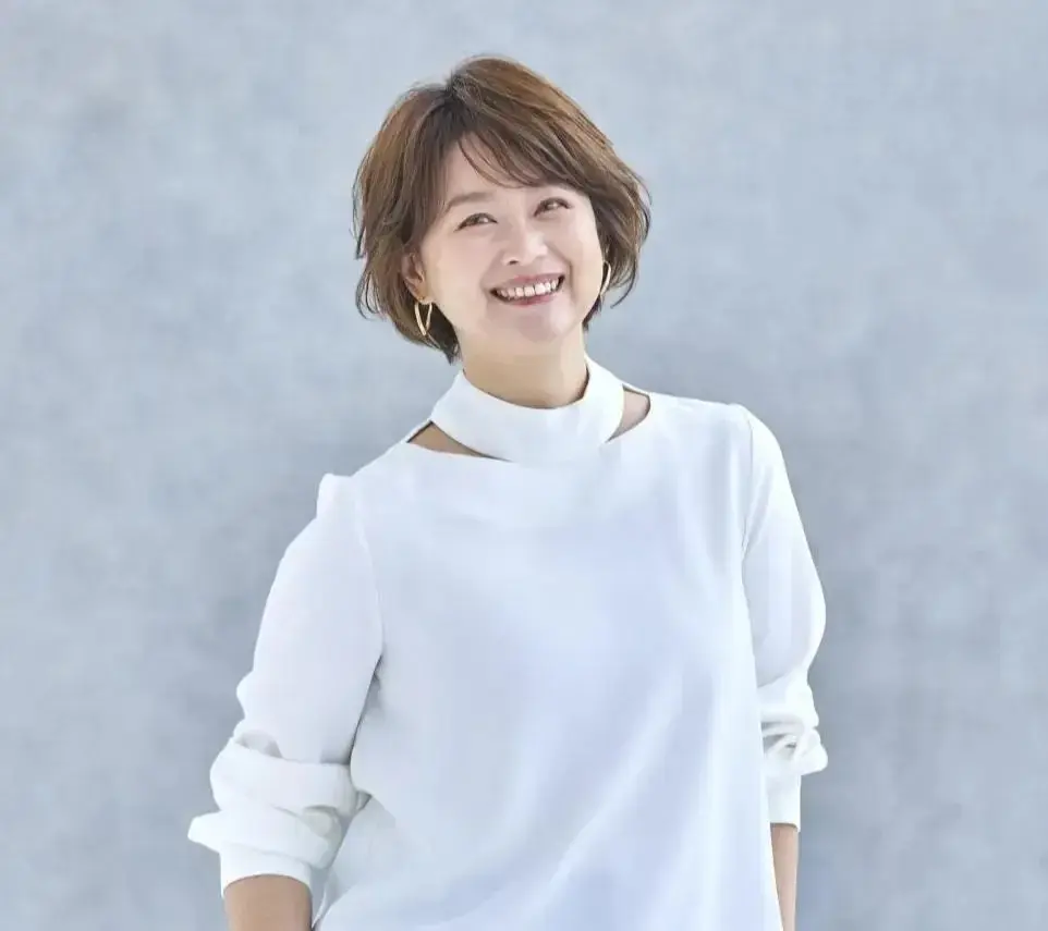 ショートヘアの女性が白いハイネックの服を着て、笑顔でカメラを見つめている。金色のイヤリングを着用。背景は無地で淡いグレー。