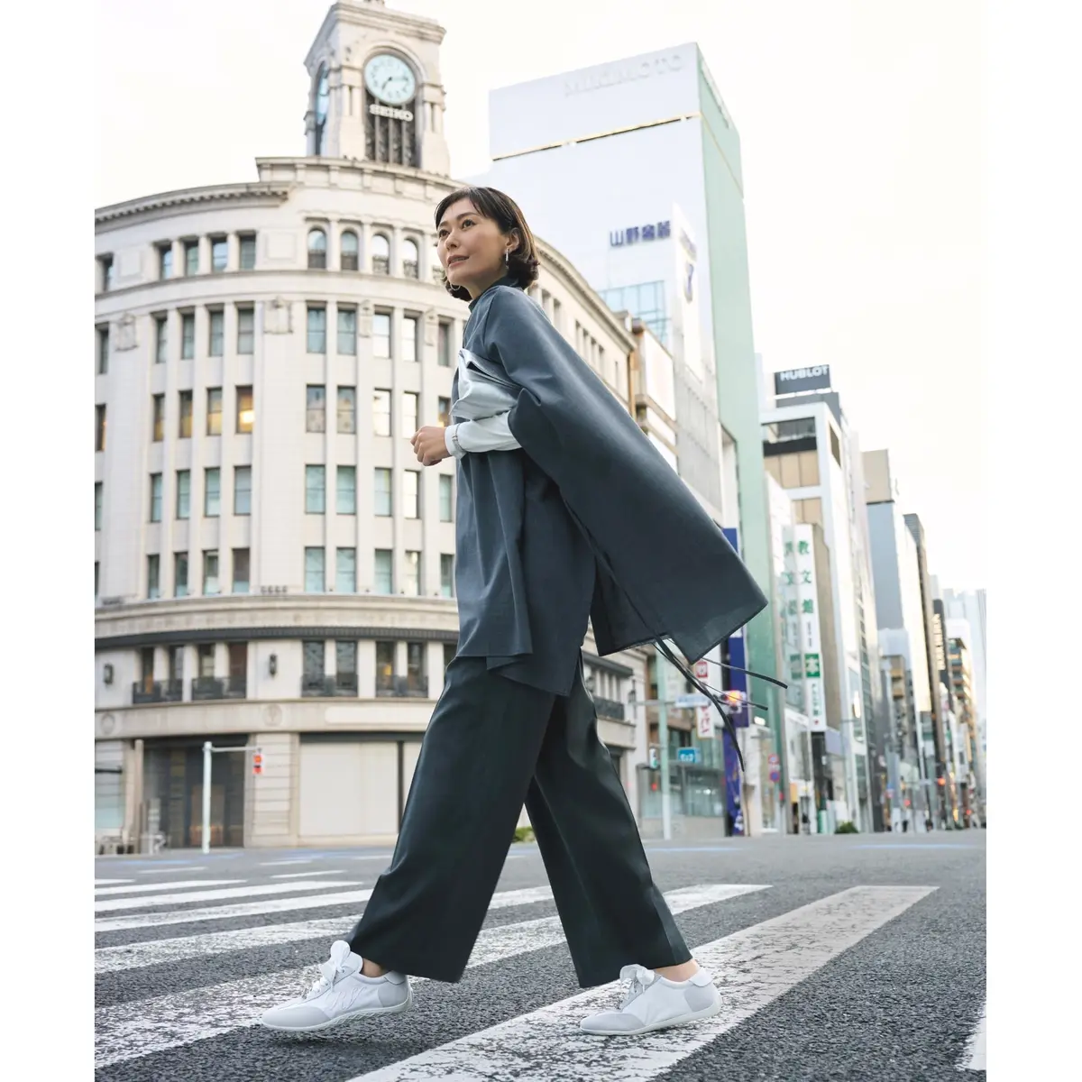 白トップスにグレーのケープアウターをはおり、ダークグレーのパンツと白スニーカーを合わせたコーディネート　モデル・田丸麻紀