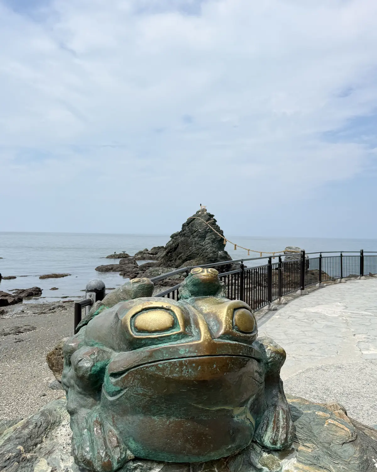 海岸に設置された青銅色の大きなカエル像。像の背中には小さなカエル像が乗り、背景には海と岩場、遊歩道が見える。空は曇り。