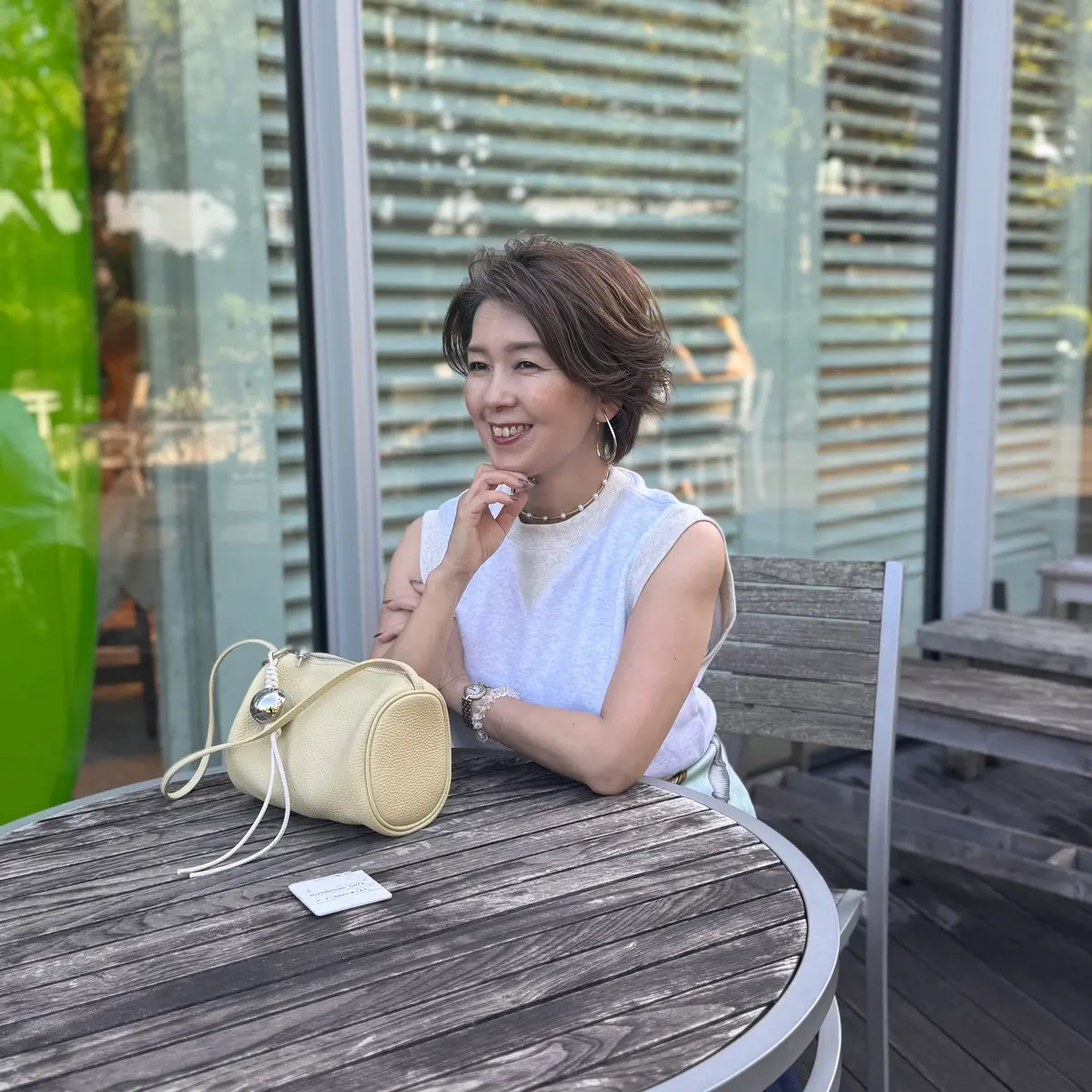 屋外のテーブルに座り、笑顔の女性。グレーのタンクトップにネックレスを着用し、右手に腕時計をしている。隣にはクリーム色のハンドバッグが置かれ、背景には緑の壁と木、窓がある。