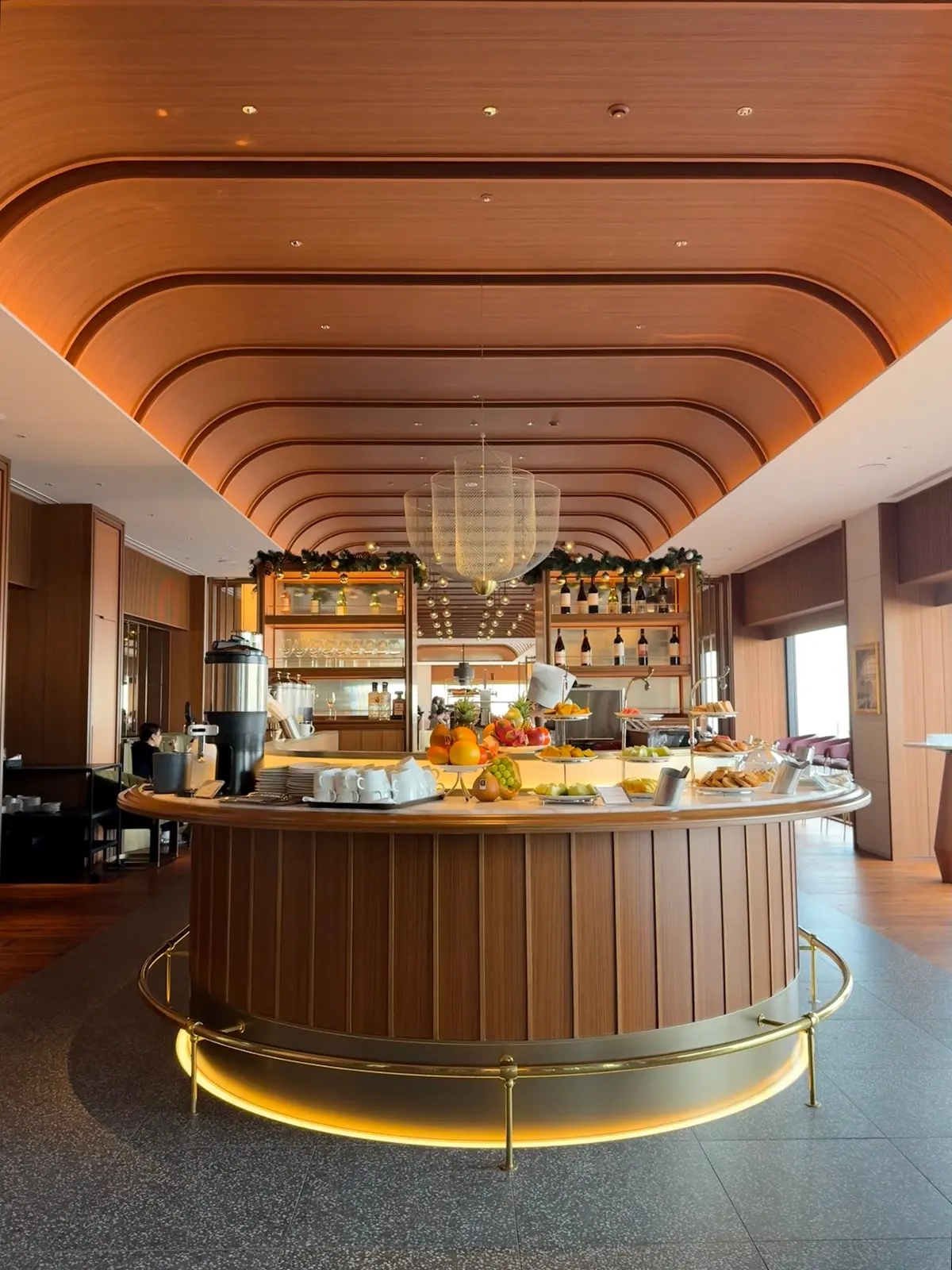 大阪　梅田　大阪ステーショングルメ　ホテルランチ　ランチビュッフェ　梅田グルメ　ホテルグルメ　丸い木製のカウンターバー、フルーツと料理の盛り合わせ、棚に並んだワインとグラス、湾曲した木製の天井とペンダントライトが特徴のモダンなバー内装