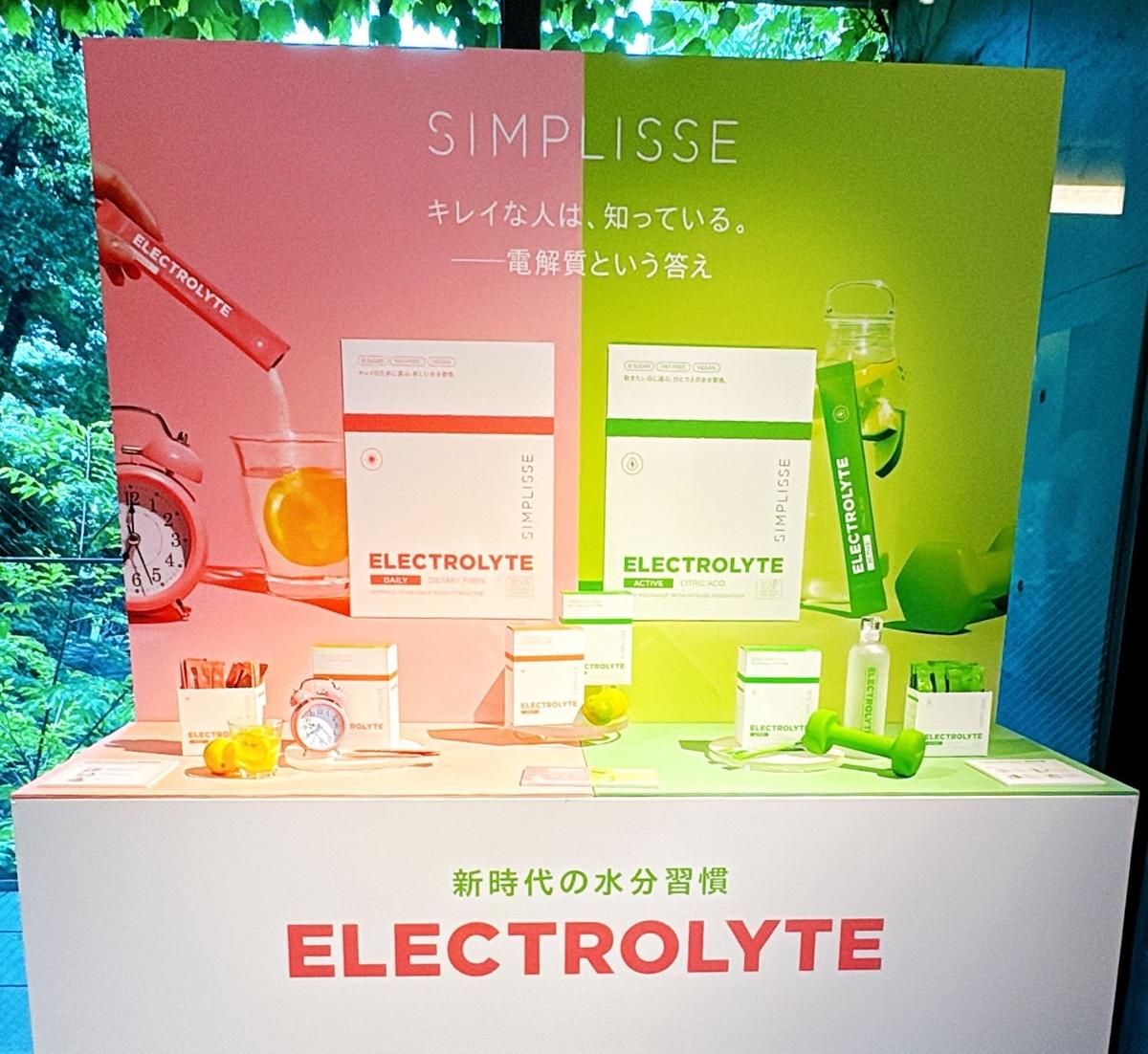 シンプリス　新作コスメ　インナーケア　エイジングケア　電解質製品の展示台に、電解質ドリンクボトル、ジュース、時計、スポーツ用具が並んでいる。背景には「SIMPLISSE」の文字と、清潔な生活を促すメッセージが書かれている。
