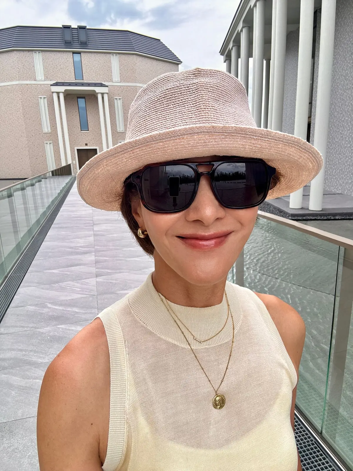 ベージュの帽子を被り、サングラスをかけた女性が、テラスで微笑んでいる。淡い黄色のトップスを着用し、ゴールドのネックレスを身につけている。背景にはモダンな建物と、曇り空が広がる。