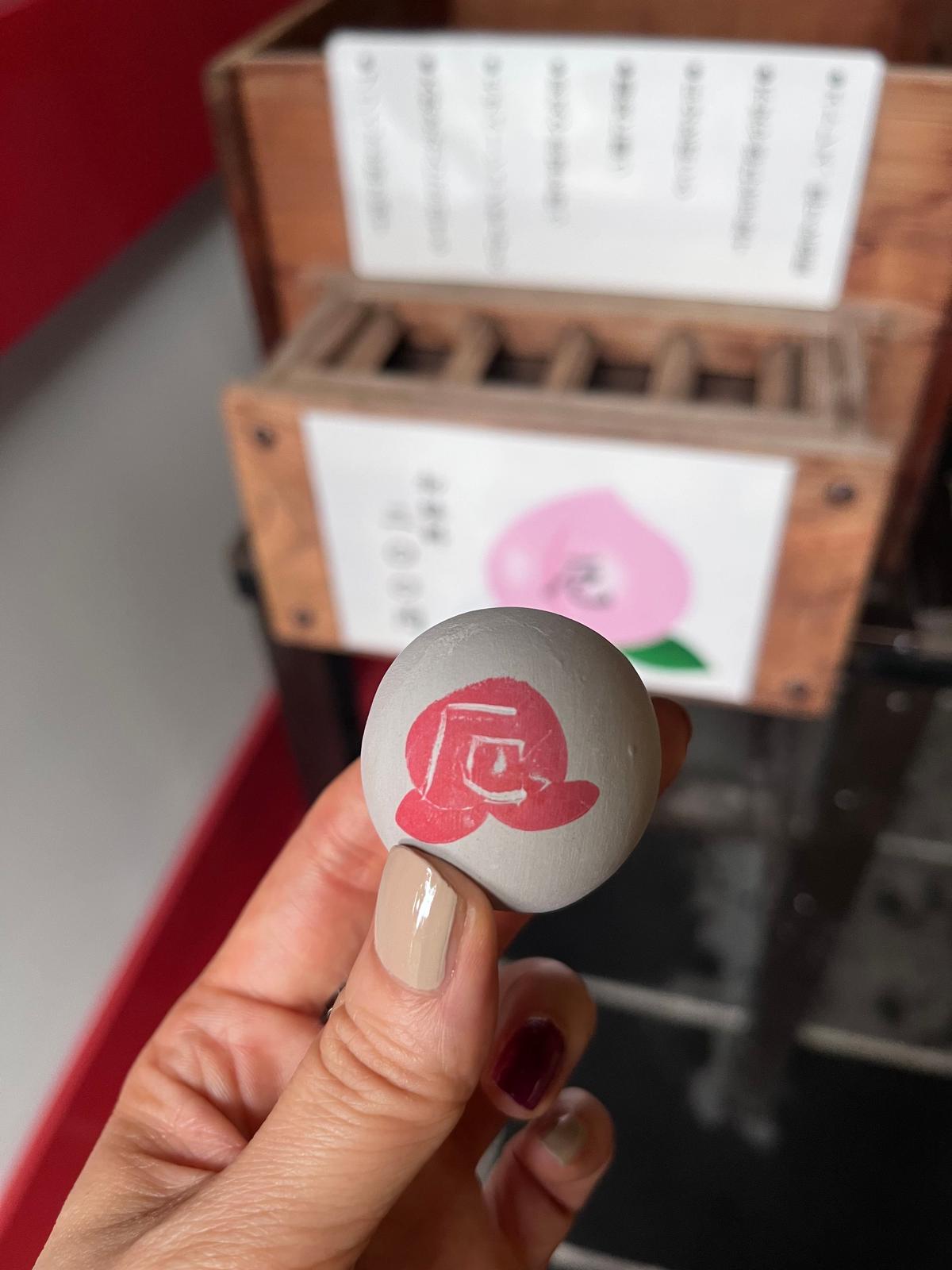 手は丸い白いボタンを持ち、赤い花のデザインが描かれている。背景には木製の箱とピンクの花のイラストが並ぶ。