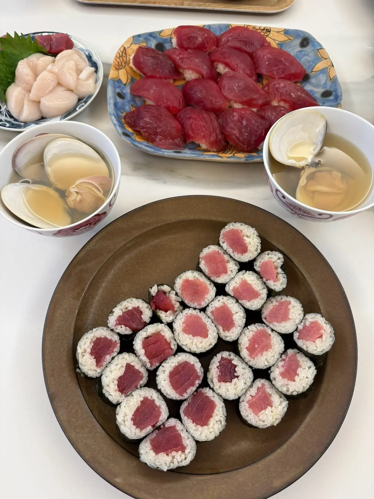 木製の皿に並んだ、マグロとご飯の巻き寿司。そばの小皿には、エビ、赤身肉、だし汁、お酒が盛られている。