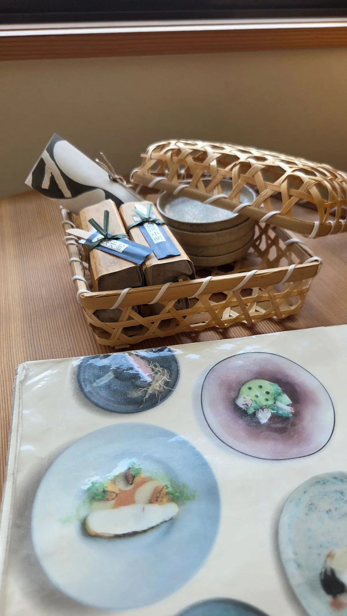 木製のテーブルに置かれた、竹製のカゴに入った小皿と、包み紙でラッピングされたお菓子。背景には、お皿のイラストが描かれた書類が置かれている。
