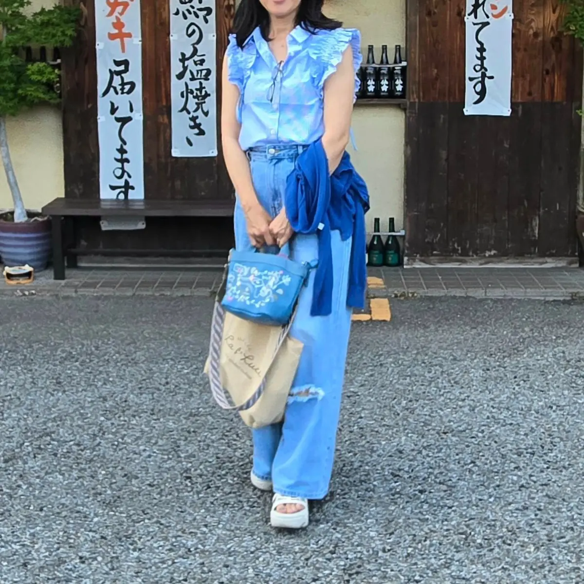 女性が青いリネンブラウスとデニムパンツを着て、木製の建物の前を歩いています。手には青いトートバッグとショルダーバッグをかけ、右手には青いカーディガンを持ち、左手には紙袋を持っています。背景には日本語の看板とボトルが並んでいます。