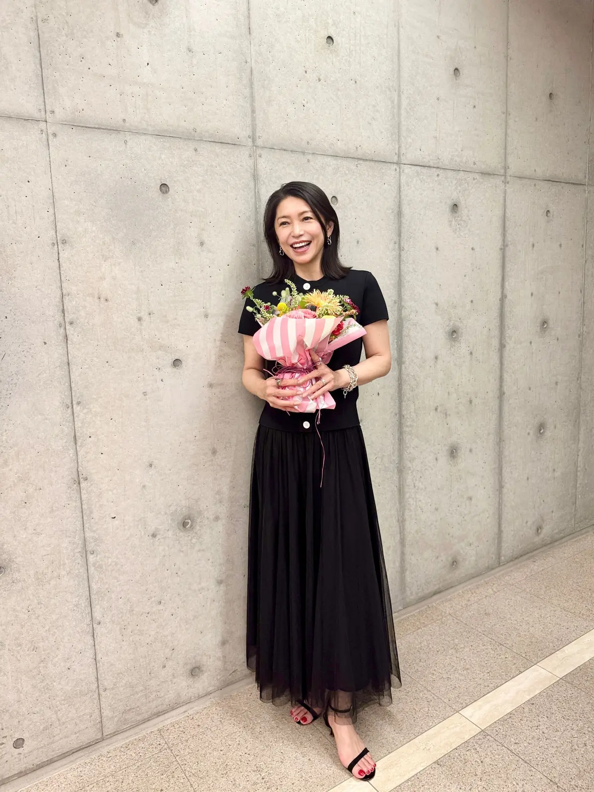 コンクリート壁の前で、花束を持つ女性。黒のロングスカートと黒のトップスを着用し、笑顔で立っている。