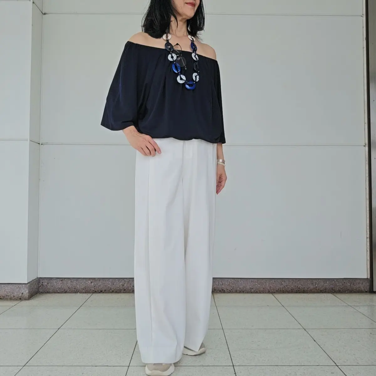 女性が黒のオフショルダートップスと白のワイドパンツを着用し、青いビーズのネックレスを身につけています。背景は白い壁と床です。
