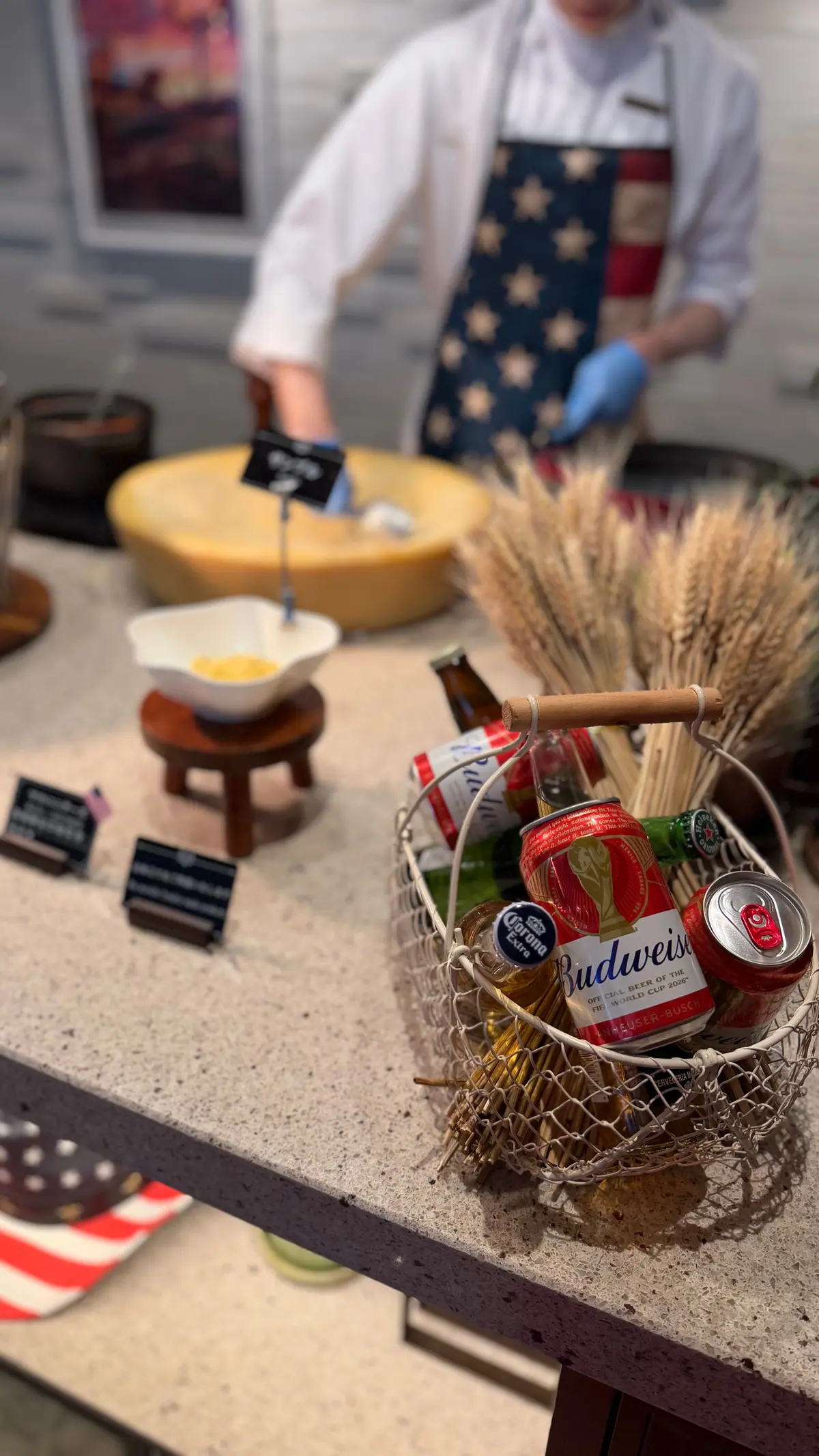 イベント会場のカウンターに置かれた、ビールやソフトドリンクが入ったワイヤーバスケット。背景には料理人が作業しており、小麦の穂も飾られている。