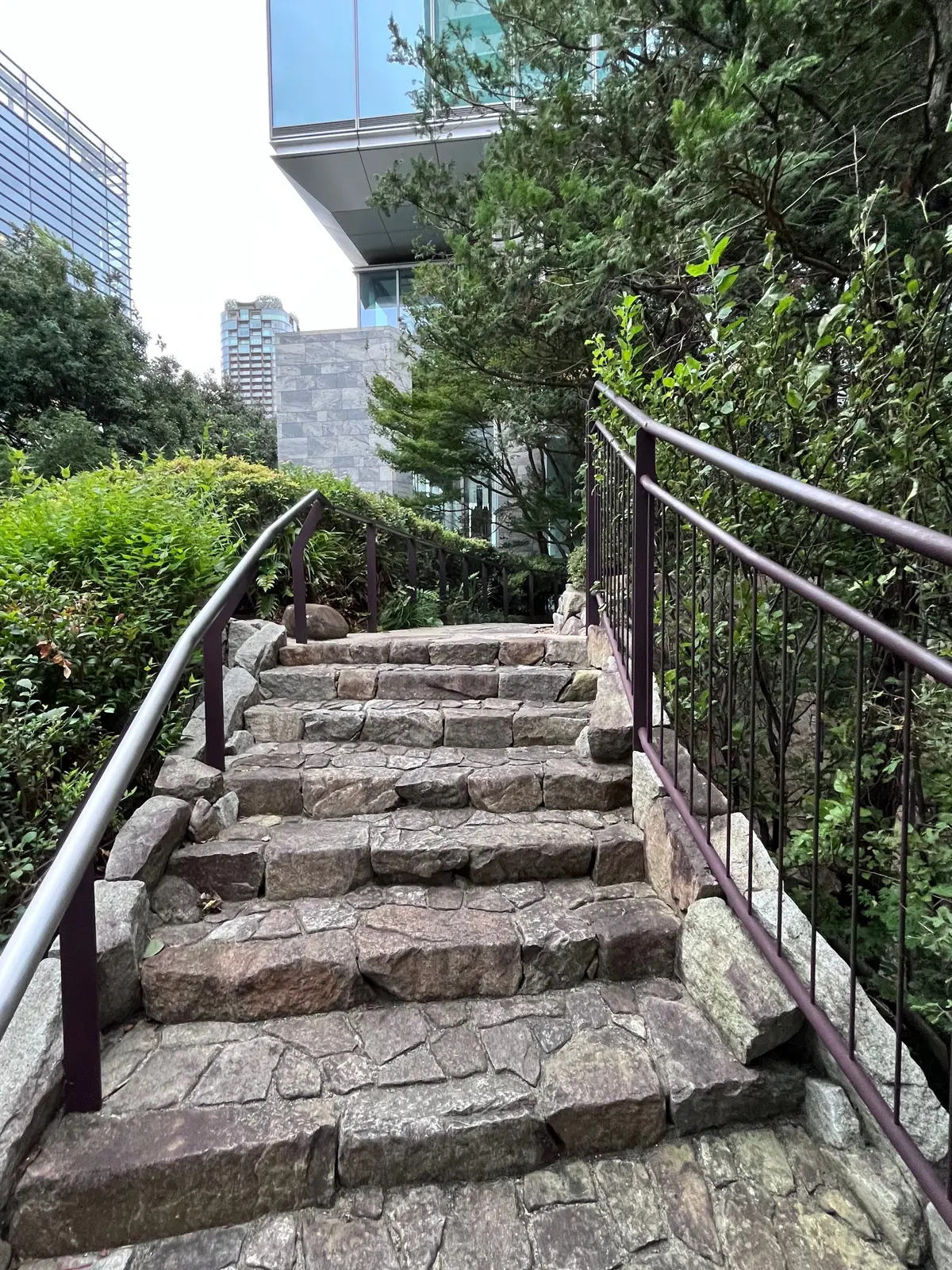 石積みの階段が、緑豊かな植栽に囲まれて上へと続いています。手すりが左右に設置され、背景には現代的な建築物が見えます。