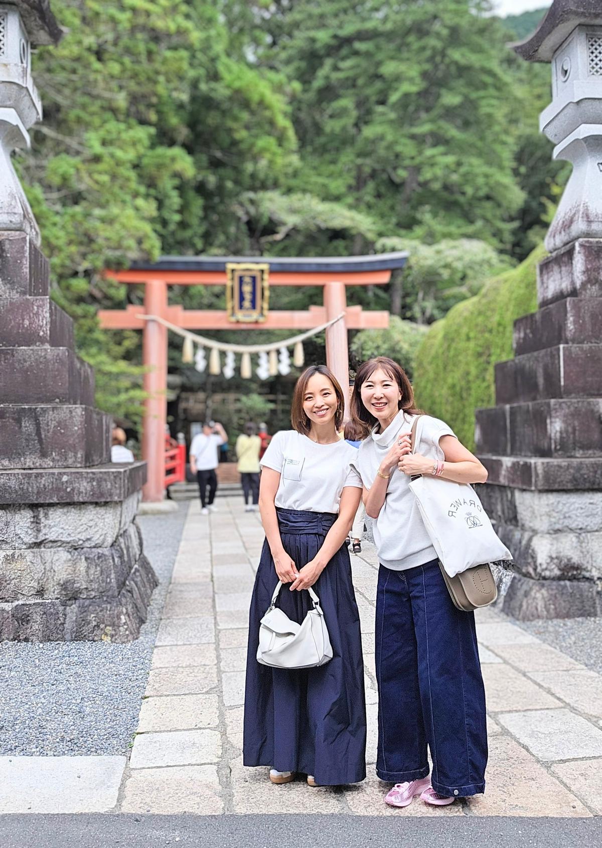 青い朱色の鳥居の前で、白いTシャツに青いスカートとジーンズを合わせた2人の女性が笑顔で立っている。右の女性は白いバッグを持ち、左の女性は白いハンドバッグを持っている。背景には緑豊かな山々が見える。