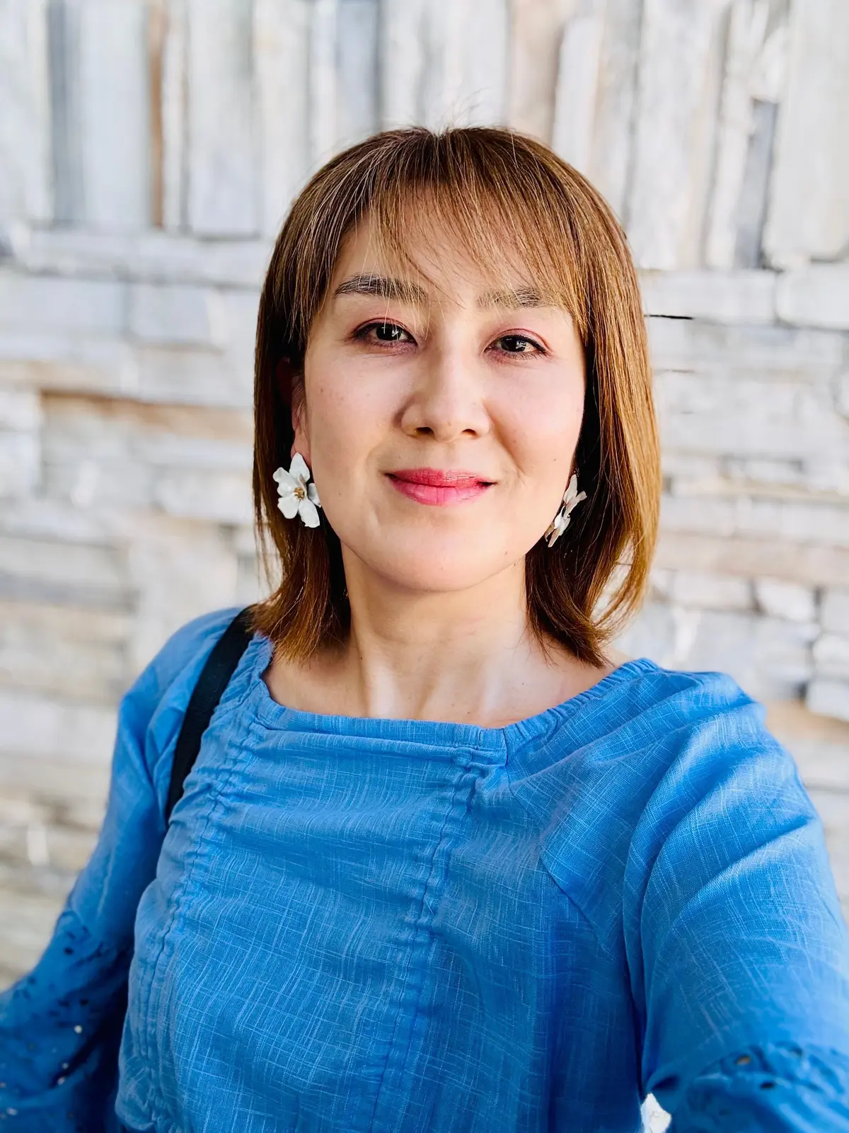 女性が青い織物製の服を着て、石造りの背景に立っています。白い花のイヤリングを着用し、ほほ笑んでいます。