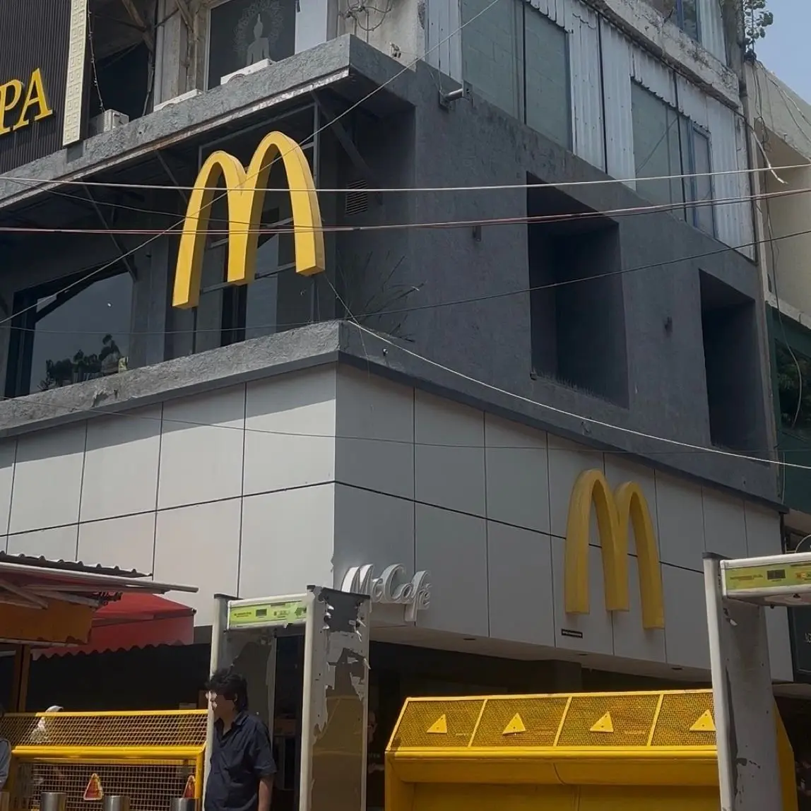 マクドナルドの店舗の外観で、黄色のMロゴが目立つ。建物はコンクリートで、窓ガラスとバルコニーがある。通りを歩く男性が写っている。