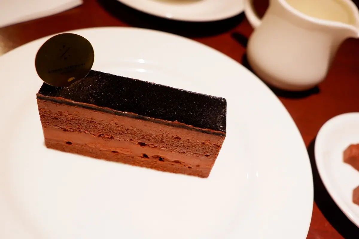 ピエール・エルメのチョコレートケーキ。濃い茶色の層に覆われた、赤茶色のムースケーキ。白い皿に盛り付けられ、金色のロゴ入りタグが添えられている。背景には白い皿とカップが写る。