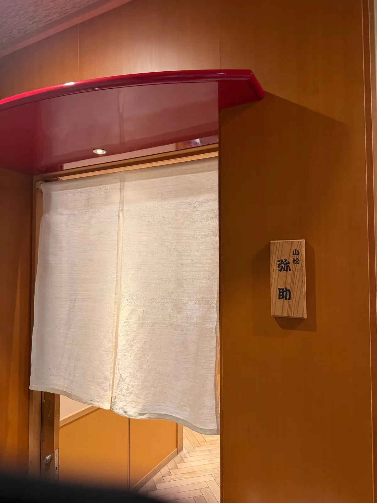 右側に木製の看板、左側には白い布製の仕切りが垂れ下がっている。背景はオレンジ色の壁。看板には黒い文字が書かれている。