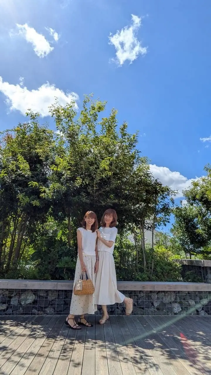 青空を背景に、白いワンピースを着た2人の女性が並んで立っている。木製のデッキに立ち、緑豊かな植物が生い茂る景色が広がる。