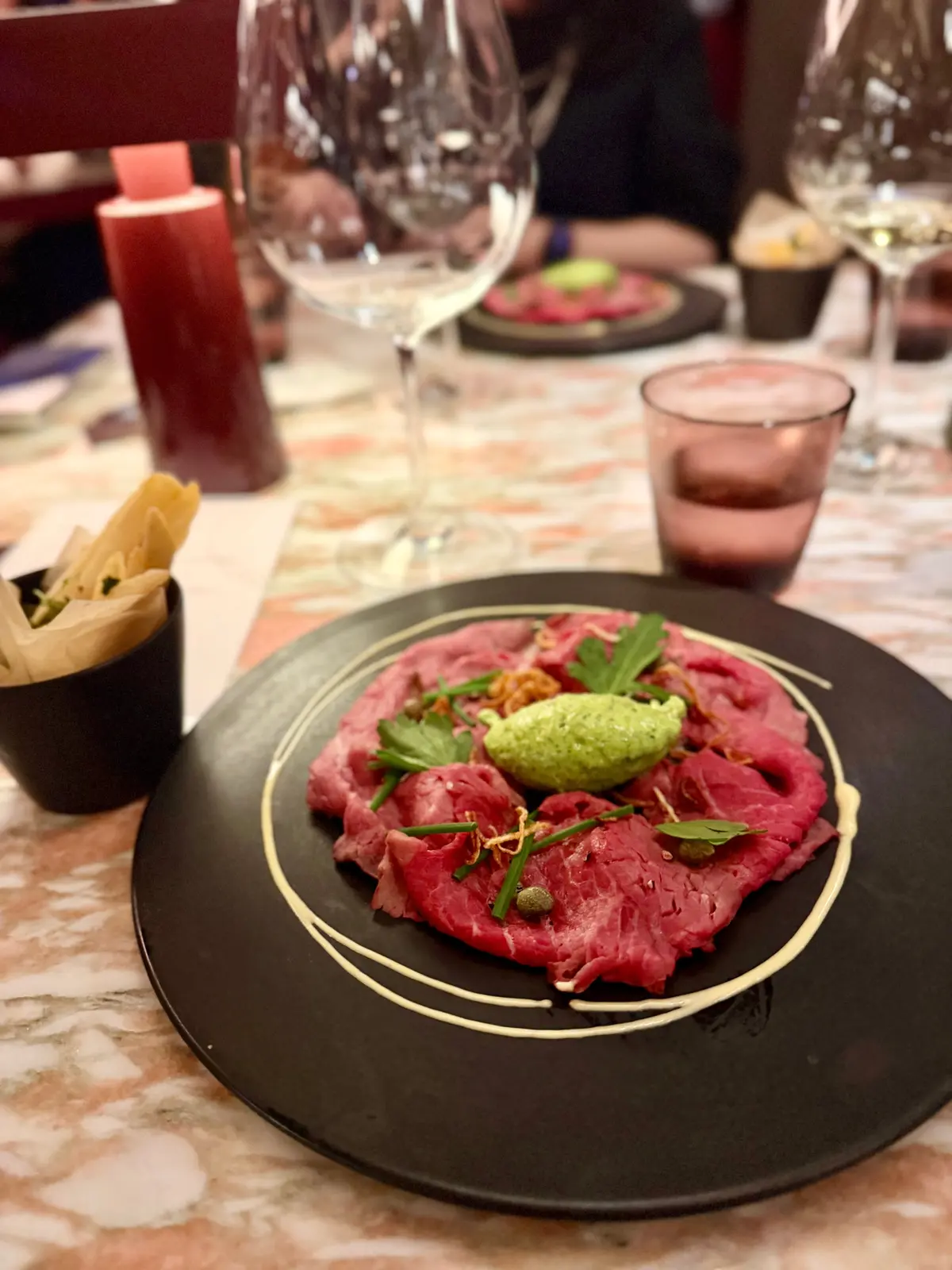 黒いプレートに赤身の牛肉が盛り付けられ、グリーンのソース、葉野菜、細い金色のラインが添えられた料理。背景にはワイングラスや他の料理が置かれている。