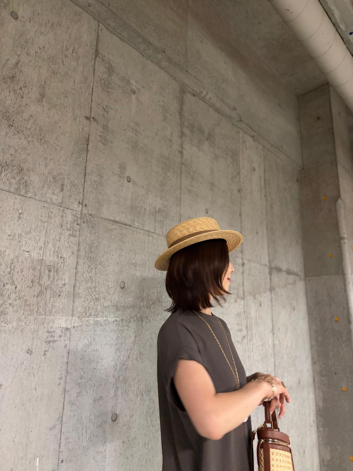 ストローハットをかぶった女性が、コンクリート壁の前で立ち、右手で小型の鞄を持っている