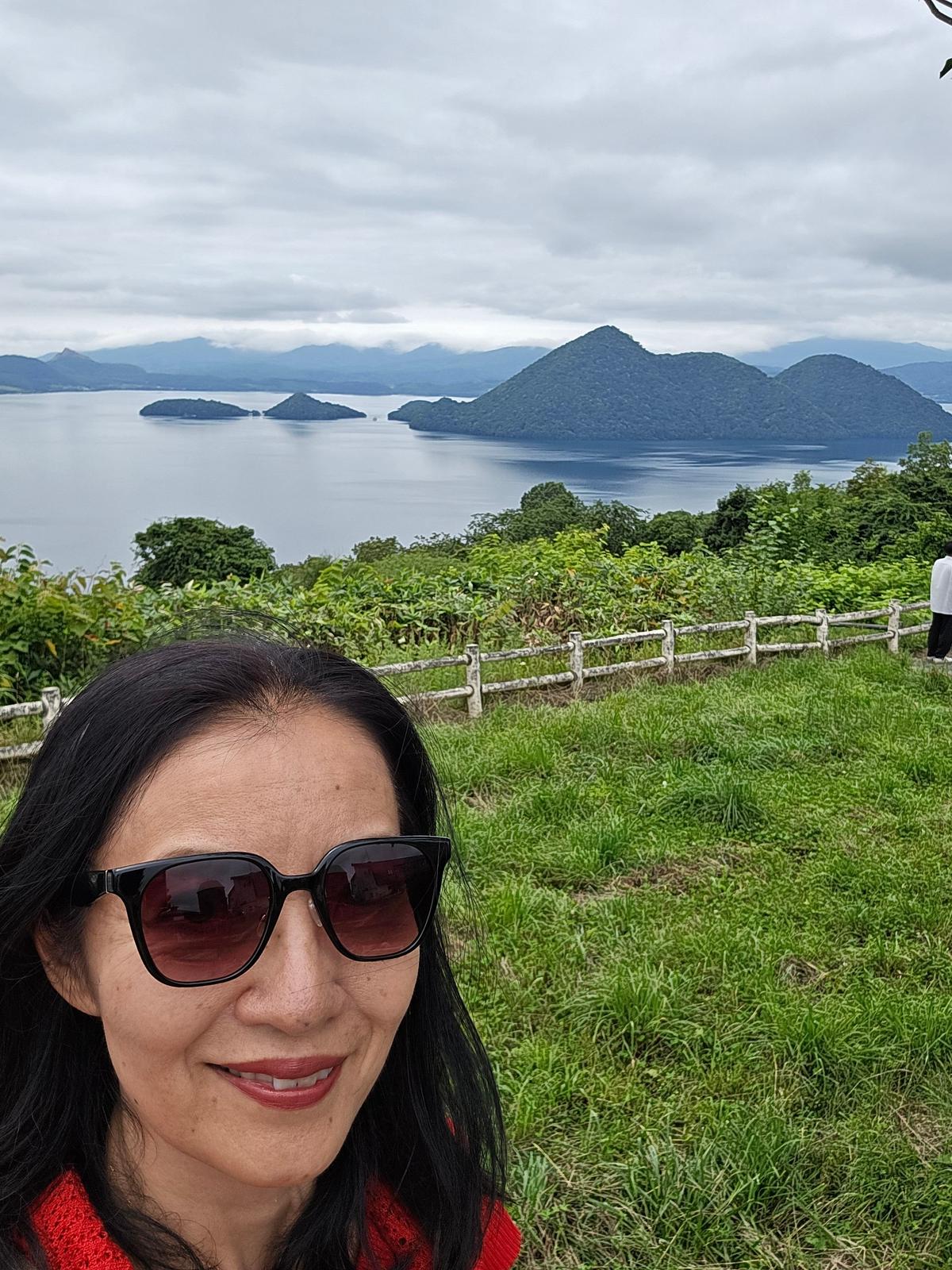 赤いセーターを着た女性がサングラスをかけ、緑の草地から湖と島々を望む風景を背景に写っている。空は曇り。