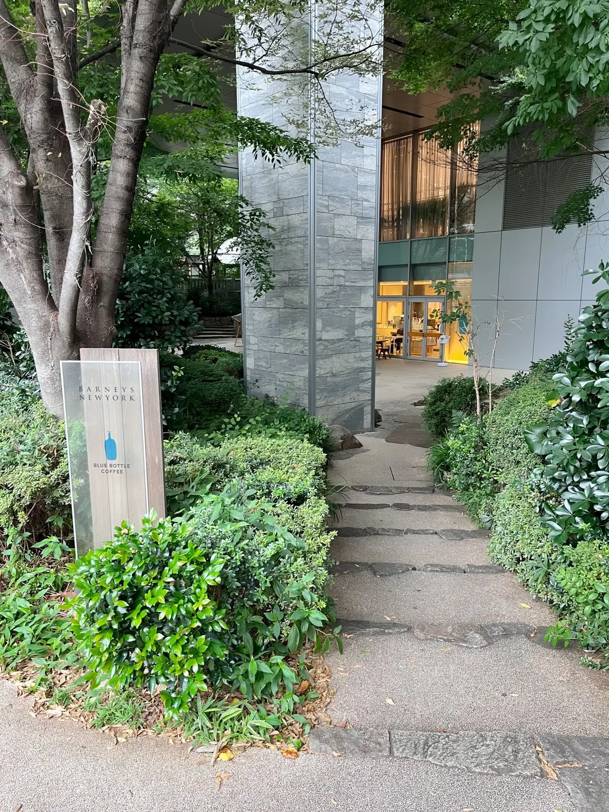 緑豊かな木々が茂る並木道。その奥には、石積みの柱と白い建物の一部が見える。手前には広いコンクリートの歩道が伸びている。