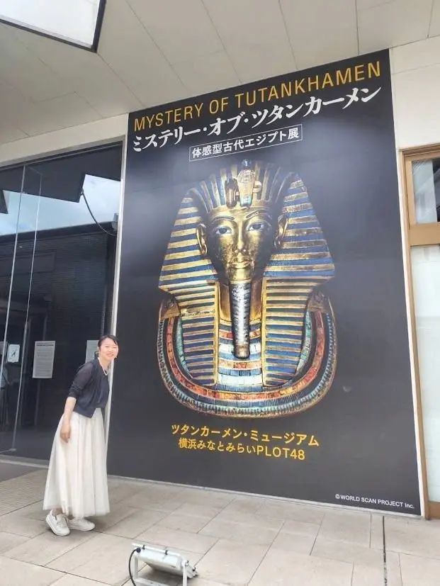 「ミステリー オブ ツタンカーメン」展のポスターの前で女性が笑顔でポーズをとっている。ポスターにはツタンカーメンの黄金のマスクが大きく描かれており、日本語のタイトルが上部に、説明文とロゴが下部に記載されている。
