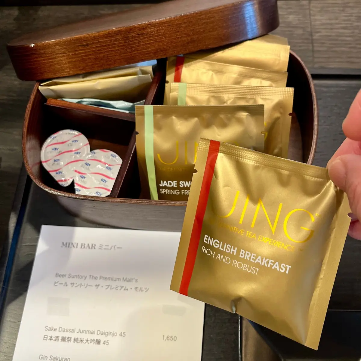 木製の箱から取り出された茶のパッケージとミニバーカードが並ぶ。茶のパッケージは金色で「JUING」と黄色の文字が入り、「ENGLISH BREAKFAST RICH AND ROBUST」と記載されている。お部屋のミニバー