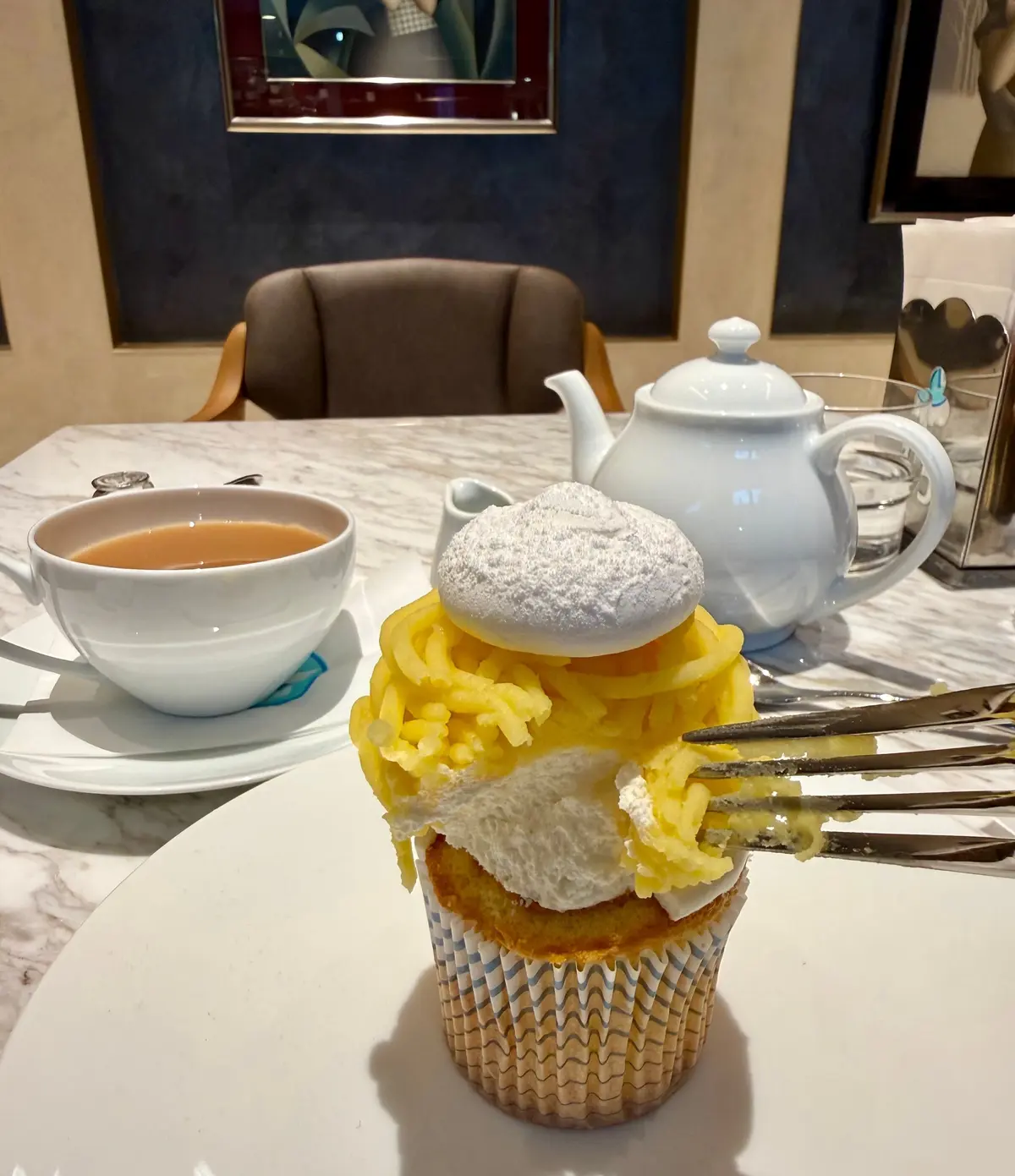 白地に青い柄のカップと茶壺、そして黄色のクリームとパウダーシュガーをかけたカップケーキが置かれたテーブル。背景には肘掛け椅子と絵画が見える。