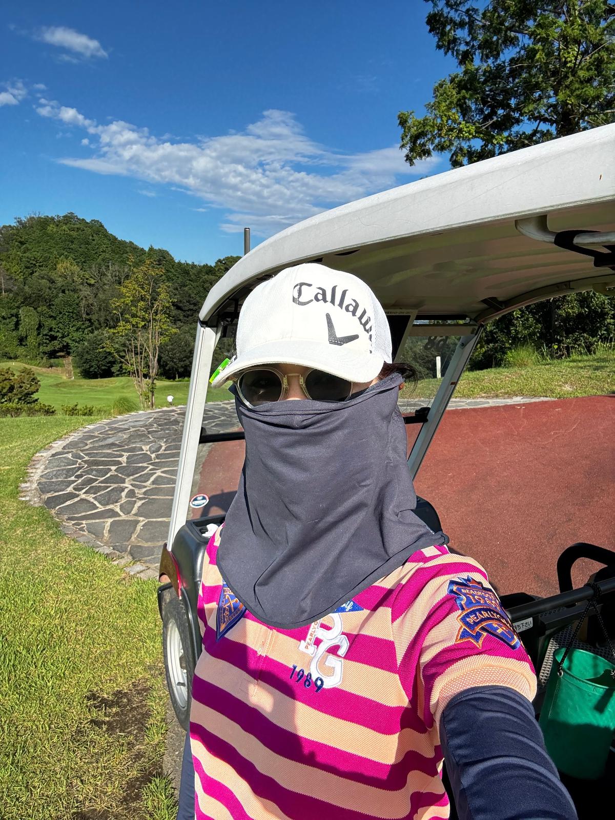 ゴルフカートに乗り、Callawayの帽子を被った男性。サングラスと黒いフェイスマスクを着用し、ピンクと白のストライプのシャツを着ている。背景には緑のゴルフコースと青空が広がる。