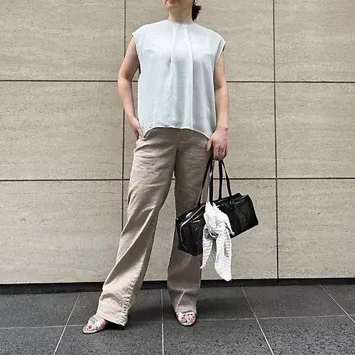 ベージュのパンツと白いトップスを着た女性が立っている写真。女性は黒いバッグを持ち、白いサンダルを履いている。背景はベージュ色のタイル壁。