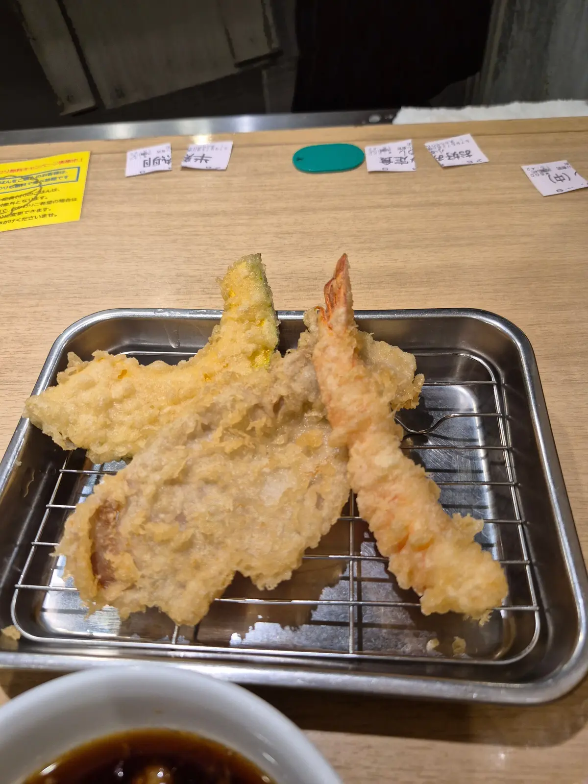 金属製のトレイに盛り付けられた天ぷら。エビ、ナス、ピーマンがそれぞれ揚げられており、薄い衣で覆われている。手前には醤油が注がれた小皿。
