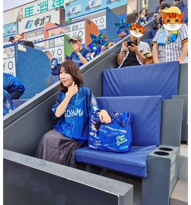 青い応援グッズを持つ女性が座席に座り、背景には同様のグッズを持つ観客がいるスタジアムの様子。